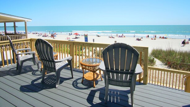 A wooden deck with chairs and a table overlooking a beach with people and umbrellas.