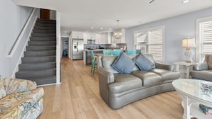 A modern living room with a grey leather sofa, a glass coffee table, and a staircase leading to a kitchen area.