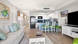 A modern living room with a sofa, coffee table, and a TV. There is a kitchen area with a fridge, microwave, and a dining table with chairs. - Beautiful ocean & beach views, oceanfront condo w/pool, sundeck, family friendly-North Myrtle Beach (North Myrtle Beach)