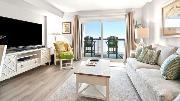 A modern living room with a flat-screen TV, a green upholstered chair, a sofa with patterned cushions, and a balcony with a view of the sea. - Beautiful ocean & beach views, oceanfront condo w/pool, sundeck, family friendly-North Myrtle Beach (North Myrtle Beach)