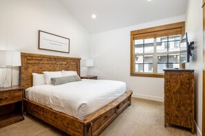 A bedroom with a wooden bed frame, white bedding, a bedside table with a lamp, a wooden cabinet, and a window with a view of buildings.