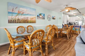 A dining area with wicker chairs and a glass table, a kitchen with white cabinets, and a ceiling fan. - Oceanfront duplex steps from beach w/shared pool, game room, kids' pool, grill (North Myrtle Beach)
