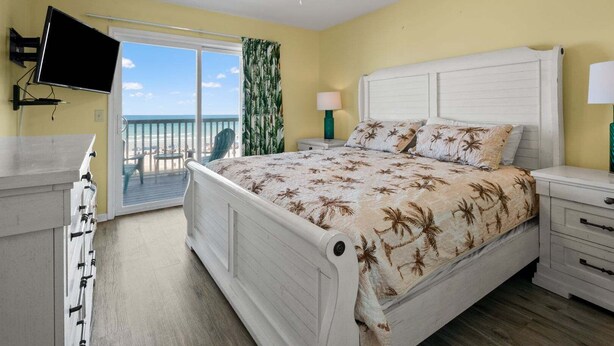 Un dormitorio con cama, televisión, balcón y vista a la playa, además de ventilador de techo.