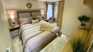 Ein Schlafzimmer mit einem großen Bett, Nachttischen, einer Lampe, einem Holz-Kopfteil, einer Wandleuchte, einer Zimmerpflanze und einem Fenster mit Vorhängen.