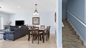 Een ruime woonkamer met een bank, eettafel en stoelen, een trap en een ingelijst kunstwerk aan de muur.