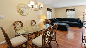 A dining area with a glass table and chairs, a chandelier, and a black sofa in the living room.