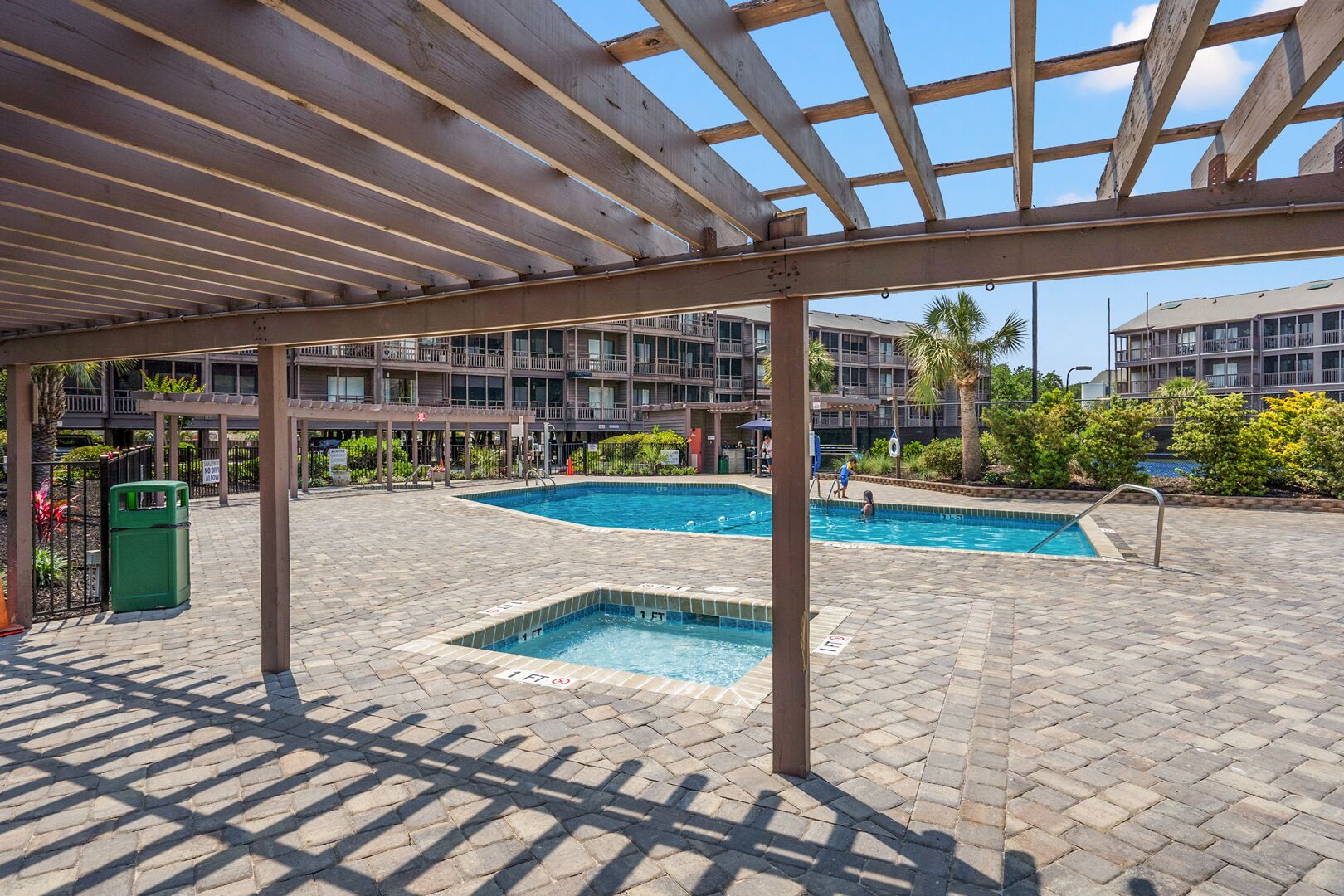 A covered outdoor swimming pool area with two pools, a pergola overhead, and a brick patio.