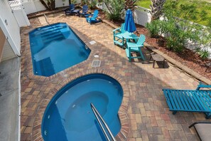 Una piscina con borde de ladrillo y dos zonas separadas, rodeada de mobiliario de patio y una pequeña mesa.