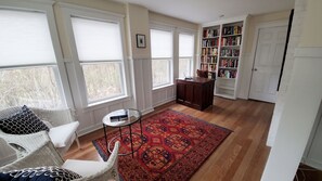 A spacious living room with large windows, a wooden floor, a red patterned rug, a bookshelf, and a white door. - Ideal Madison home just steps to the beach (Madison)