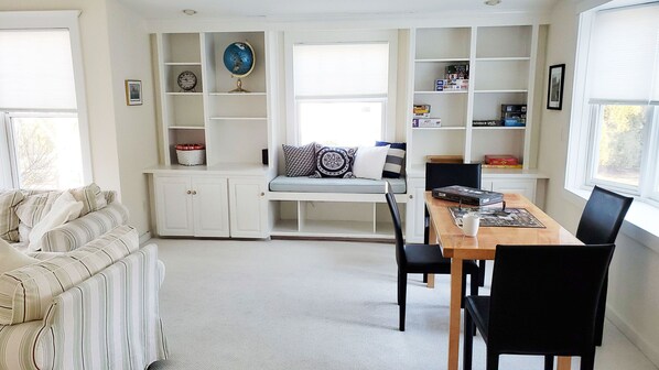 A cozy living room with a sofa, a dining table with chairs, a built-in bookshelf, and a window with blinds. - Ideal Madison home just steps to the beach (Madison)