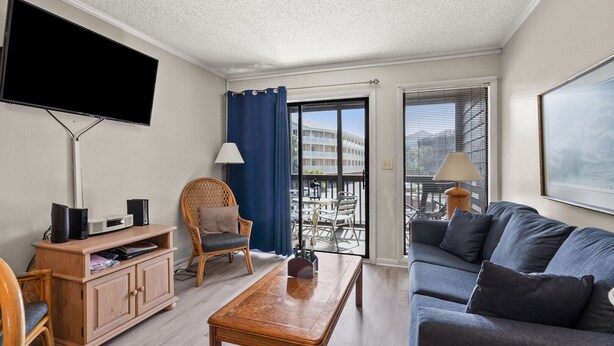 A modern living room with a flat-screen TV, wooden furniture, a sofa, and a balcony with a view of buildings. - Upscale family resort w/all the amenities-large pool, tennis courts, hot tub and more (North Myrtle Beach)