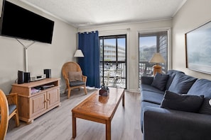 A modern living room with a flat-screen TV, wooden furniture, a sofa, and a balcony with a view of buildings.