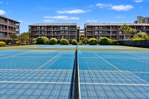 A tennis court with a net in the center, surrounded by buildings with balconies and greenery. - Upscale family resort w/all the amenities-large pool, tennis courts, hot tub and more (North Myrtle Beach)