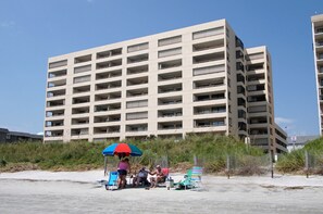 A multi-story building with numerous balconies and a sandy ground area with beach chairs and an umbrella.
