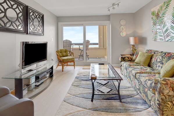 A living room with a TV, a glass coffee table, a sofa, and a chair. There is a balcony with a view of the ocean.
