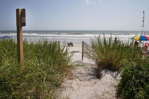 A sandy beach path leading to the ocean, with a wooden signpost and a beach umbrella in the background.