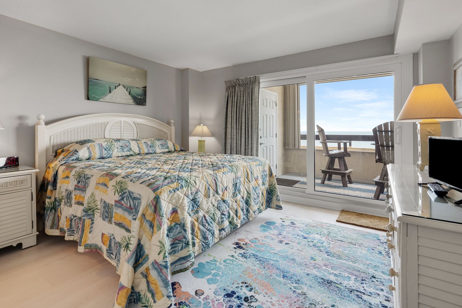 A bedroom with a bed, a desk with a computer, a chair, a window with a view of the beach, and a door leading to a balcony.