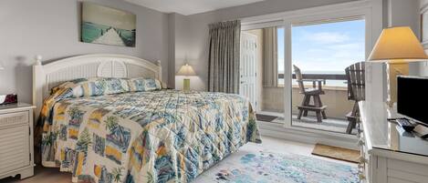 A bedroom with a bed, a desk with a computer, a chair, a window with a view of the beach, and a door leading to a balcony.