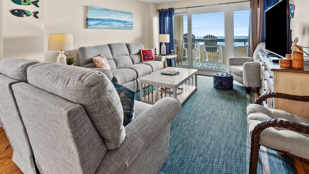 A modern living room with a grey sofa, a small table, and a television. There is a balcony with a view of the sea. - Step into luxury at SEA POINTE 802, where your personal beachfront paradise awaits. (North Myrtle Beach)