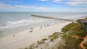 A beach with a pier extending into the water, sandy beach, and a few umbrellas. - Step into luxury at SEA POINTE 802, where your personal beachfront paradise awaits. (North Myrtle Beach)