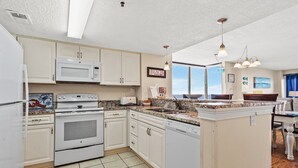 A kitchen with white cabinets, a microwave, stove, and dishwasher. There is a window with a view of the ocean and a sign that says "BEACH". - Step into luxury at SEA POINTE 802, where your personal beachfront paradise awaits. (North Myrtle Beach)