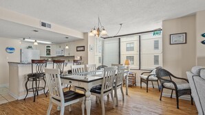 A spacious dining area with a wooden table and white chairs, a kitchen with white cabinets, and large windows with blinds. - Step into luxury at SEA POINTE 802, where your personal beachfront paradise awaits. (North Myrtle Beach)