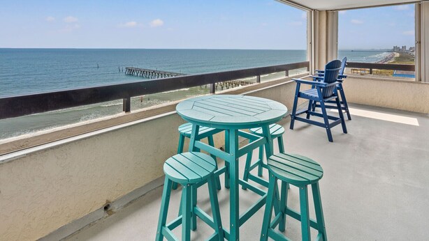 A balcony with a view of the ocean, featuring two green tables and blue chairs.