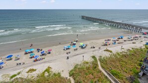 A beach with a pier extending into the ocean, people relaxing under umbrellas, and a path leading to the beach.