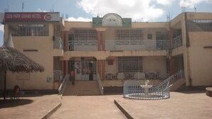 A multi-story building with a sign for the Gem Park Grand Hotel. There are stairs leading to the entrance, a thatched roof structure to the left, and a fenced area with a circular object in the center. - Gempark Grand Hotel (Thika)