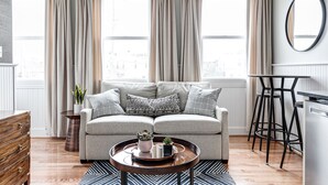 A modern living room with a grey sofa, a round wooden coffee table, and a view through large windows with beige curtains. - Brand New, Gorgeous Studio Close to Everything! (Richmond)