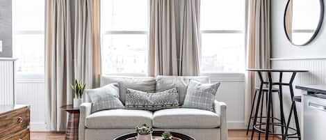 A modern living room with a grey sofa, a round wooden coffee table, and a view through large windows with beige curtains.