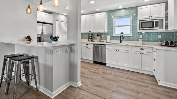 A modern kitchen with white cabinets, a stainless steel dishwasher, a microwave, and a window with teal backsplash. - Channel home w/gorgeous saltwater pool, relaxing porches-fish, swim, sunbathe, enjoy! (North Myrtle Beach)
