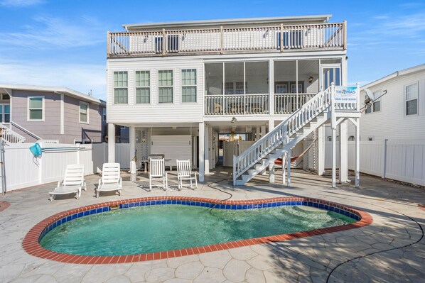Uma casa de dois andares com varanda, um alpendre com cadeiras e uma piscina.