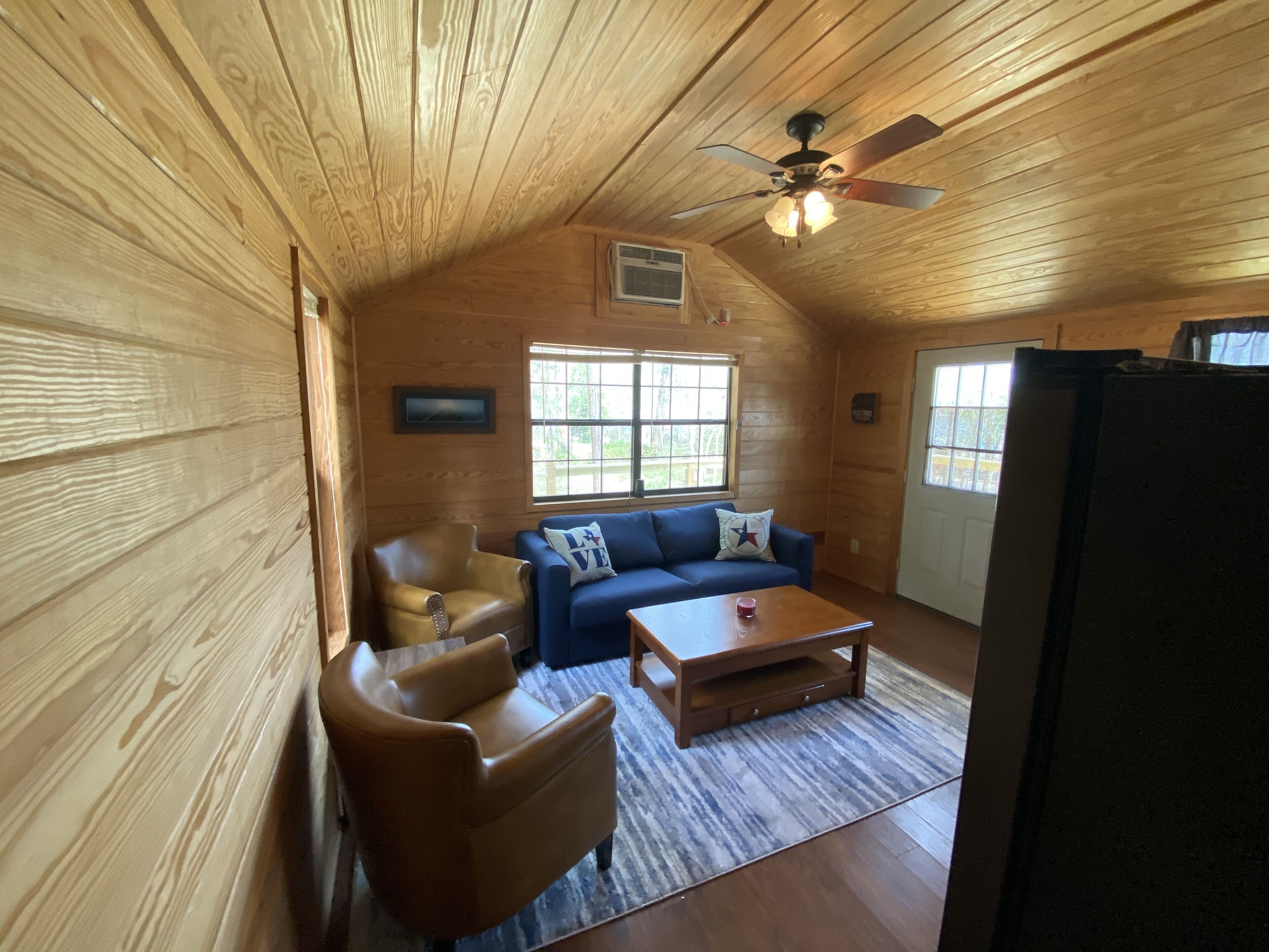 L’intérieur d’un chalet accueillant, avec un plafond en bois, un ventilateur de plafond, un canapé bleu, un fauteuil en cuir marron, une table basse en bois et un réfrigérateur.