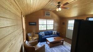 Interior acogedor de una cabaña con techo de madera, un ventilador de techo, un sofá azul, un sillón de cuero marrón, una mesa de centro de madera y un refrigerador.