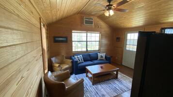 L’intérieur d’un chalet accueillant, avec un plafond en bois, un ventilateur de plafond, un canapé bleu, un fauteuil en cuir marron, une table basse en bois et un réfrigérateur.