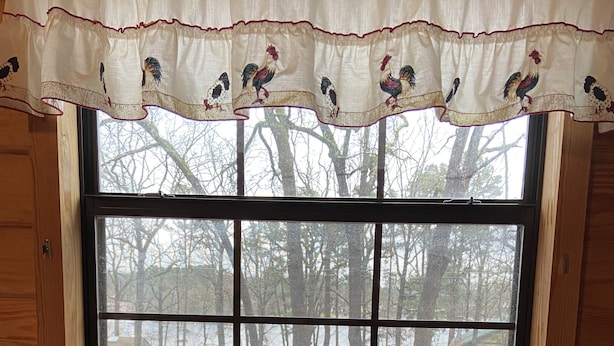 Una cocina con un lavamanos doble de acero inoxidable, un ventanal con un valance decorativo, y vistas a árboles y una casa desde el exterior.