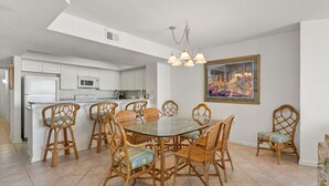 A spacious dining area with a glass-top table surrounded by wicker chairs, a kitchen in the background, and a painting on the wall. - Perfect for families or groups looking for an upscale beachfront experience. (North Myrtle Beach)