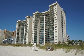 A high-rise hotel with multiple balconies, located near a beach with a lifeguard stand and a beachside sign. - Classy oceanfront family-sized condo rental w/lazy river, pool, and fitness center near Ocean Drive (North Myrtle Beach)