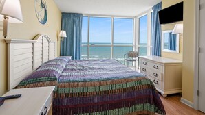 Ein Schlafzimmer mit einem großen Bett, einem Schreibtisch, einem Stuhl, einer Kommode und einem Fernseher. Das Zimmer bietet Blick auf den Ozean.
