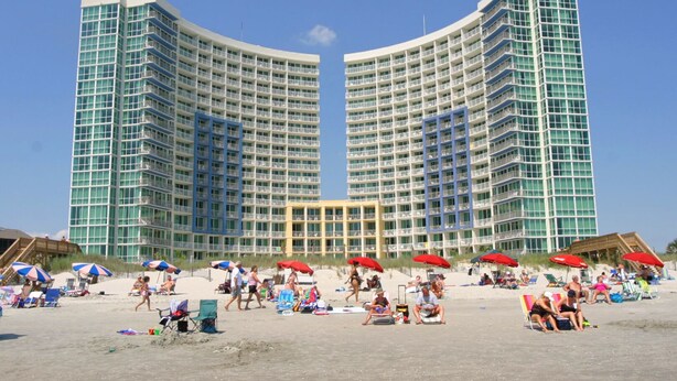 Ein großes Hotel mit mehreren Balkonen, einem Sandstrand davor und einigen Strandsonnenschirmen.