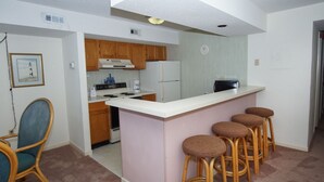 A compact kitchen with wooden cabinets, a white refrigerator, a stove, and a bar area with stools. - Spacious oceanfront condos w/pool and sundeck-walking distance to Cherry Grove Pier (North Myrtle Beach)