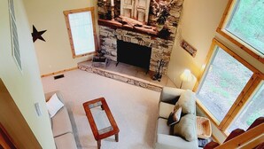 Ein gemütliches Wohnzimmer mit Kamin aus Stein, einer Holztreppe und Blick ins Freie durch große Fenster.