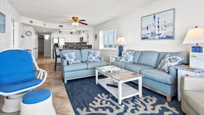 Un salon avec un canapé bleu, une table basse et un fauteuil bleu. On y trouve un ventilateur de plafond, un tableau représentant des voiliers et une lampe sur une table d’appoint.