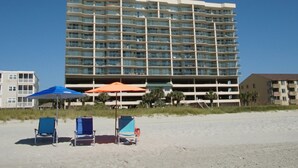 Un hôtel situé en bord de mer, doté de plusieurs étages, de balcons et de nombreux équipements, notamment des parasols et des chaises longues.