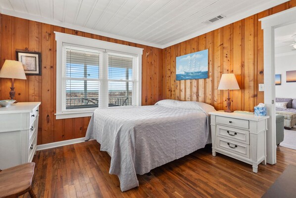 A bedroom with wooden paneling, a large window with blinds, a bed with a gray bedspread, two bedside tables with lamps, and a painting on the wall.
