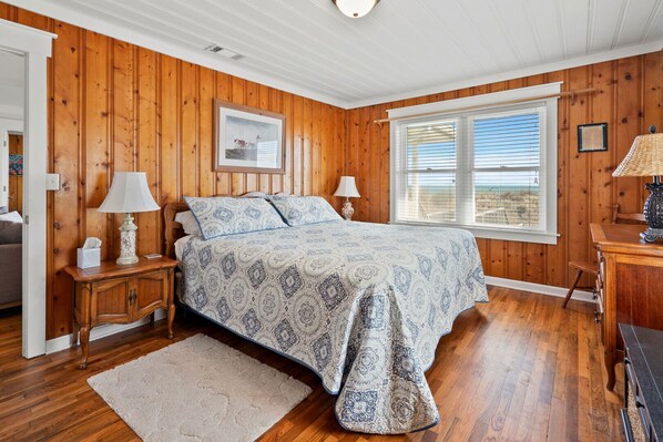 A bedroom with a wooden paneled wall, a large bed with patterned bedding, two bedside lamps, a framed picture on the wall, and a window with blinds.