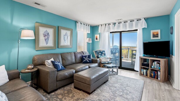 Ein Wohnzimmer mit Ecksofa, einem kleinen Tisch, einem Sessel, einer TV-Konsole mit Büchern und Blick ins Freie durch eine Schiebetür.