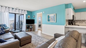 Ein Wohnzimmer mit einer Couch, Hocker und einem Fernseher. Es gibt eine Küchenzeile mit Schränken und einer Arbeitsplatte.