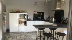 A modern kitchen with white cabinets, a built-in oven, a microwave, and a coffee maker. There are three black bar stools and a large island with a black countertop. - Architect House Near Lyon Eurexpo Stade Lumières (Saint-Priest)